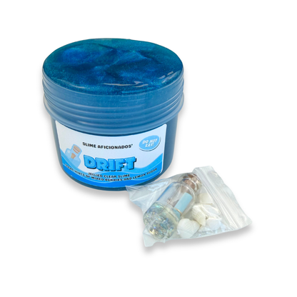 A blue jar of Drift Slime with a white and blue label, shown next to a small clear plastic bag containing white sea shells and a small cork bottle resin charm,