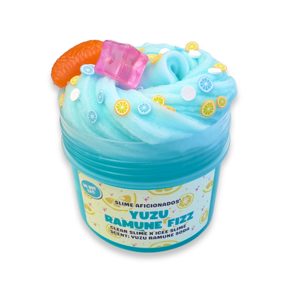 A jar of Yuzu Ramune Fizz slime, featuring light blue and turquoise swirled slime topped with a pink ice cube, a realistic 3D orange segment charm, and lemon clay slice decorations.