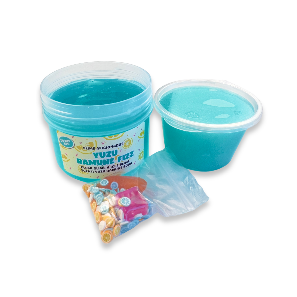A jar of bright blue clear slime next to a small plastic cup of the icee slime, with a small bag of colorful decorative slime charms (ice and orange) and clay slices.