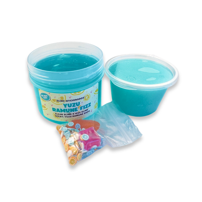 A jar of bright blue clear slime next to a small plastic cup of the icee slime, with a small bag of colorful decorative slime charms (ice and orange) and clay slices.