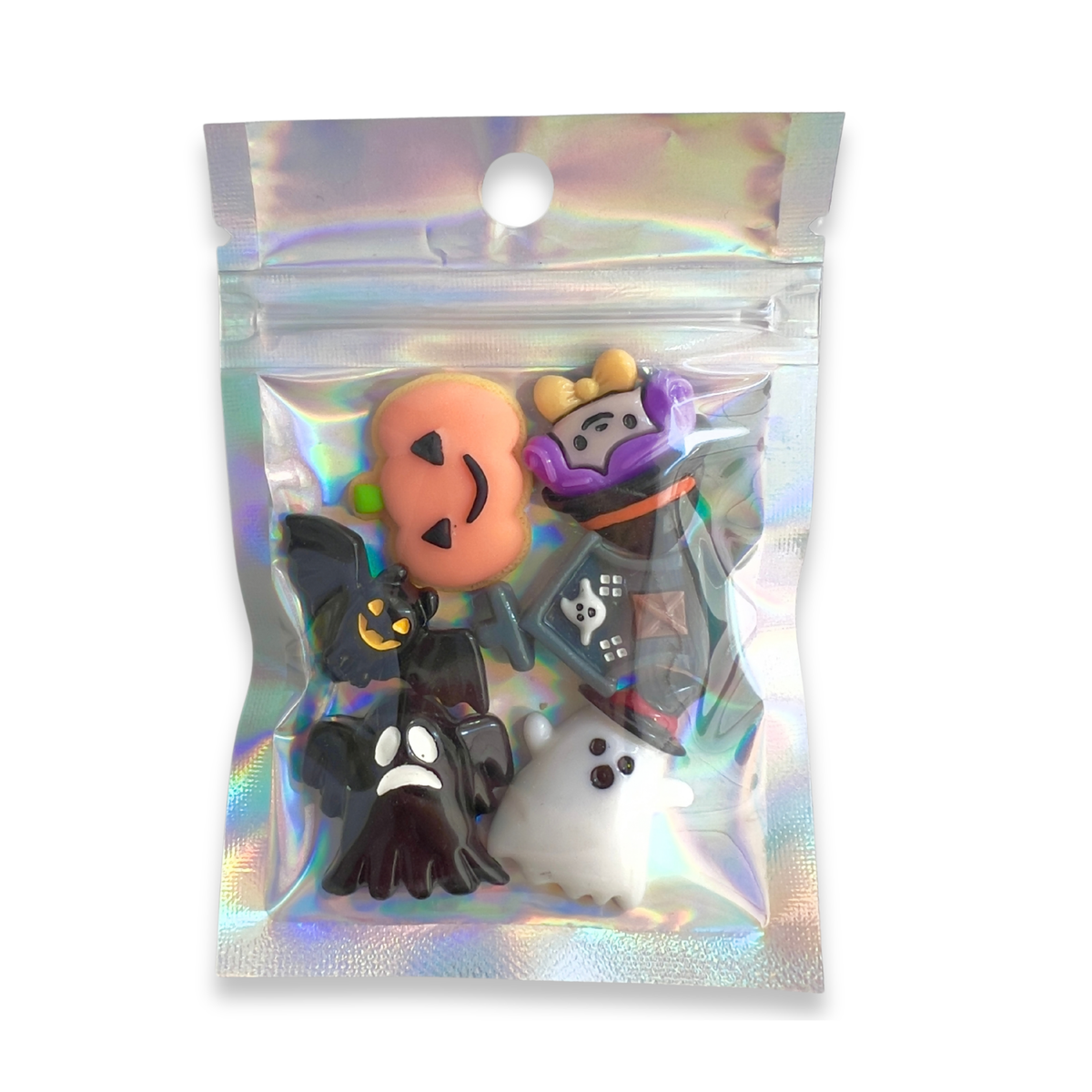A small holographic zip-top pouch containing five Halloween-themed charms or beads: a peach jack-o'-lantern, a purple wizard hat, a black bat, a white ghost, and a black skull-and-crossbones flag.
